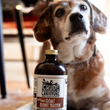 Load image into Gallery viewer, Dog sitting next to a bottle of Crude Carnivore goat bone broth on a wooden floor.